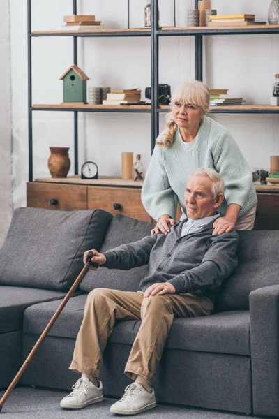 Дружина у відставці обіймає сумного старшого чоловіка, який сидить на дивані у вітальні — стокове фото
