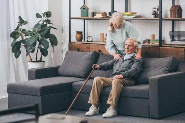 Дружина у відставці обіймає сумного старшого чоловіка, який сидить з прогулянковою тростиною у вітальні — стокове фото