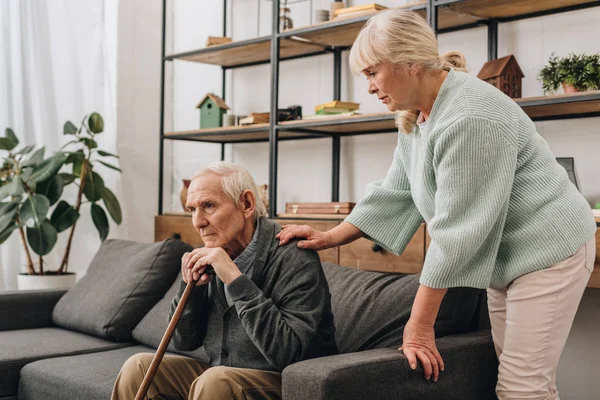 Moglie anziana in piedi vicino al marito in pensione seduto sul divano — Foto stock