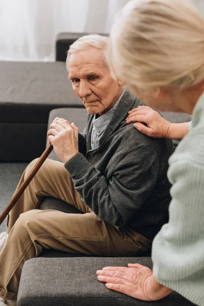 Triste marido jubilado con bastón de caminar sentado en la sala de estar cerca de la esposa mayor - foto de stock