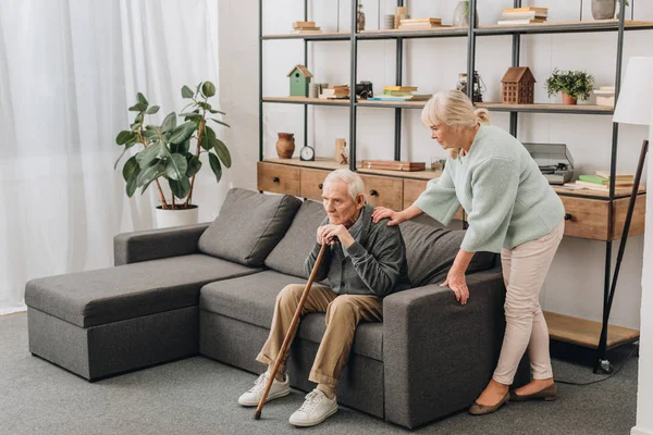 Жена на пенсии, стоящая рядом с грустным старшим мужем, сидящим с тростью на диване — стоковое фото