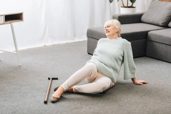Femme âgée avec des cheveux blonds couchés sur le sol près du canapé et ressentant de la douleur — Photo de stock