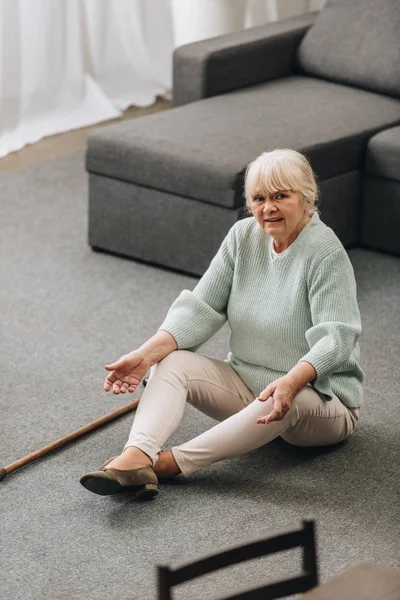 Одинокая пожилая женщина с светлыми волосами сидит на полу с тростью рядом с диваном — стоковое фото