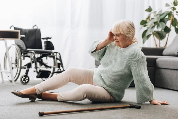 Helpless retired woman sitting on floor near wheelchair and having headache — Stock Photo