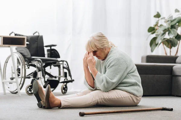Upset retired woman sitting on floor near wheelchair and holding head — Stock Photo