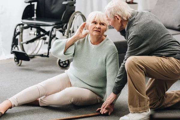 Unterstützender pensionierter Ehemann schaut Seniorin auf dem Fußboden sitzend an — Stockfoto