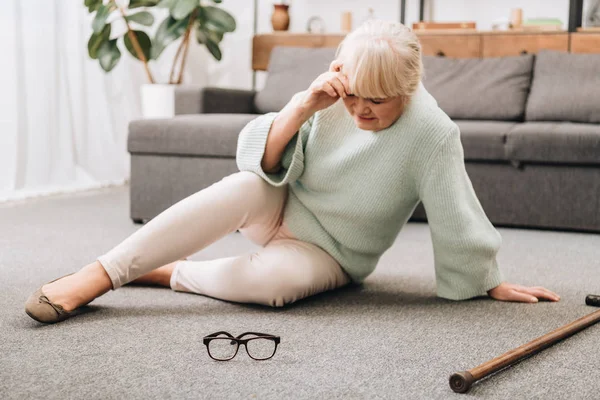 Donna in pensione seduta sul pavimento vicino a occhiali e bastone da passeggio e avendo mal di testa — Foto stock