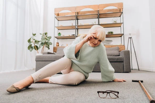Rentnerin mit blonden Haaren sitzt im Wohnzimmer auf dem Boden und hat Kopfschmerzen — Stockfoto