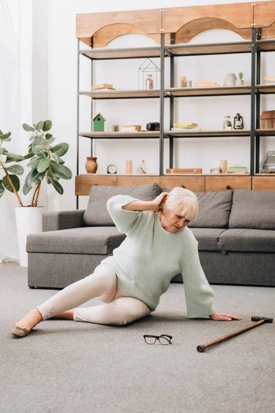 Mulher aposentada com cabelo loiro sentado no chão perto de bengala e óculos na sala de estar — Fotografia de Stock