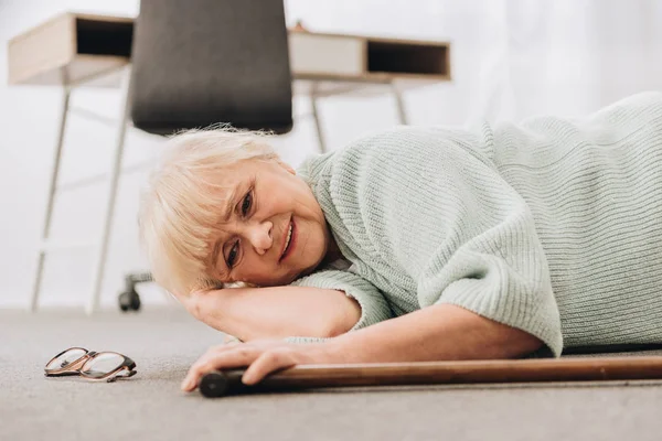 Mujer jubilada indefensa con el pelo rubio tumbado en el suelo en la sala de estar - foto de stock