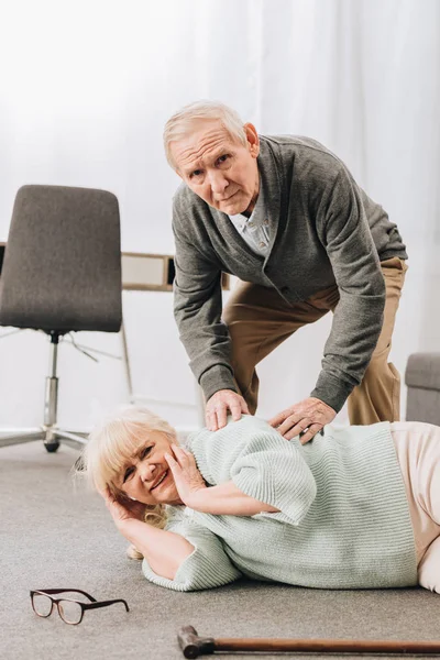 Unterstützender pensionierter Ehemann schaut Seniorin an, die am Boden liegt und Kopfschmerzen hat — Stockfoto