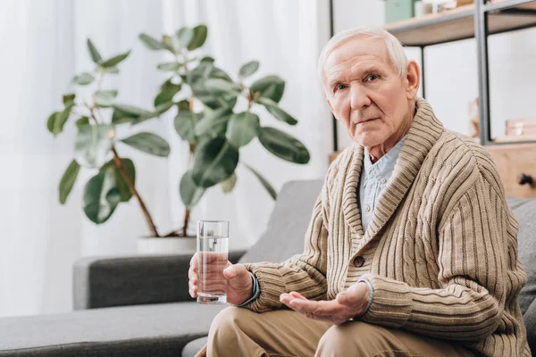 Hombre mayor sosteniendo píldoras y vaso de agua y mirando a la cámara - foto de stock