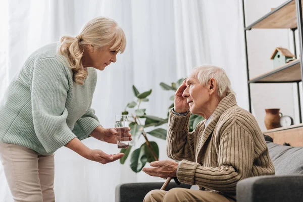 Старша жінка дає таблетки і склянку води старому чоловікові з ходьбою палицею — стокове фото