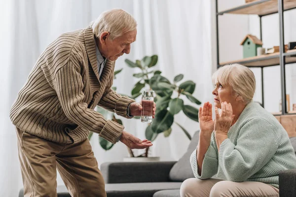Hombre mayor mirando a la vieja esposa y dando vaso de agua y pastillas - foto de stock