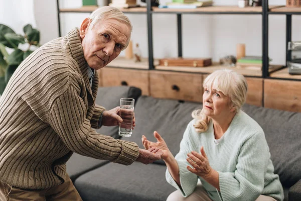 Senior man giving to old woman glass of water and pills while looking at camera — Stock Photo