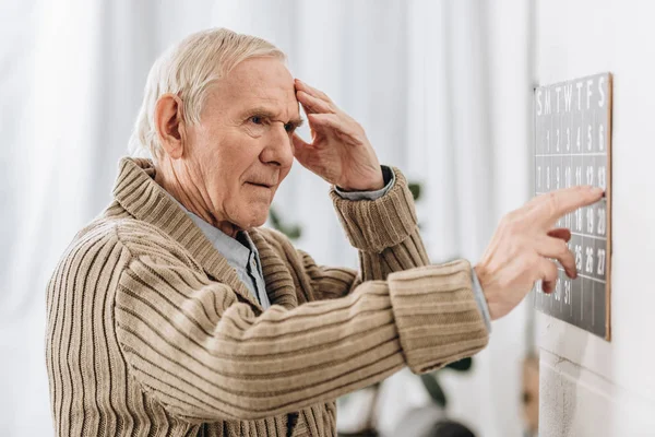 Hombre mayor mirando el calendario de la pared y tocando la cabeza - foto de stock