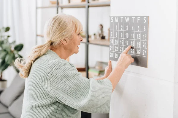Mujer mayor tocando calendario de pared y mirando las fechas - foto de stock