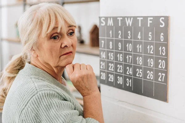 Upset senior woman touching wall calendar and remembering dates — Stock Photo
