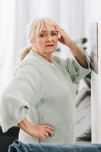 Upset senior woman standing near calendar on wall and holding head — Stock Photo