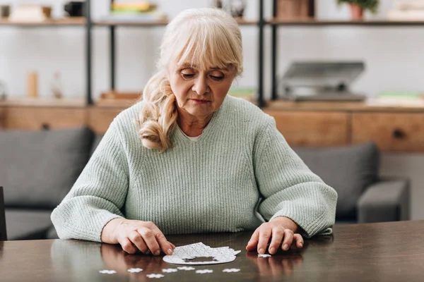 Старша жінка дивиться на шматочки головоломки на столі — стокове фото