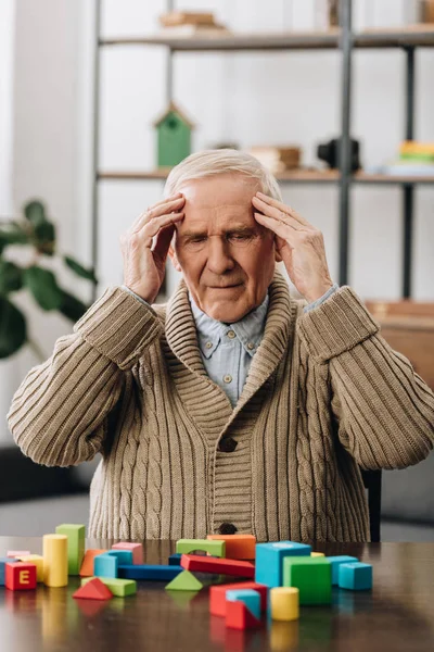 Pensionista tendo dor de cabeça enquanto sentado perto de brinquedos de madeira em casa — Fotografia de Stock