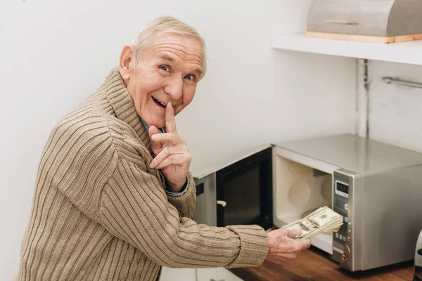 Alegre hombre mayor colocando el dedo en los labios para decir silencio mientras se pone dinero en el horno de microondas - foto de stock