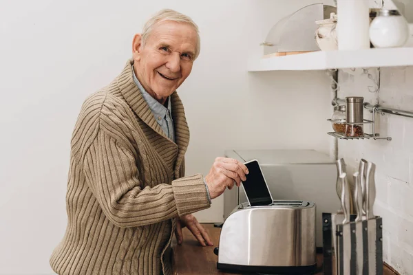 Happy senior man with dementia disease putting smartphone in toaster — Stock Photo