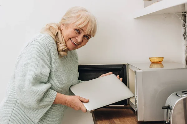 Donna anziana allegra con malattia di demenza mettendo il computer portatile in forno a microonde — Foto stock