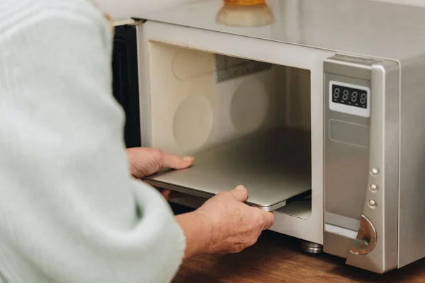 Visão cortada de mulher idosa com doença de demência colocando laptop no forno de microondas — Fotografia de Stock