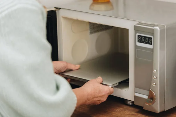 Vista ritagliata di donna anziana con malattia di demenza mettendo il computer portatile nel forno a microonde — Foto stock