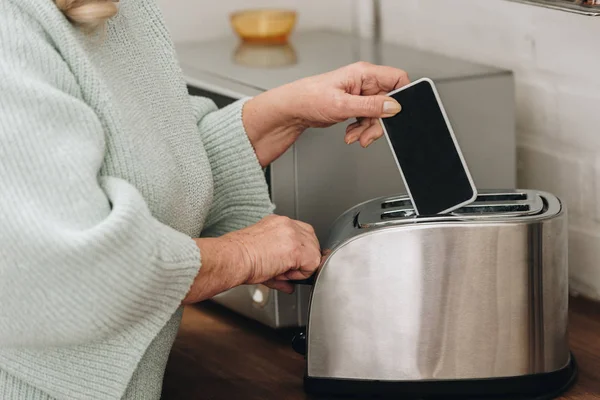 Ausgeschnittene Ansicht einer an Demenz erkrankten Rentnerin, die Smartphone mit leerem Bildschirm in Toaster legt — Stockfoto