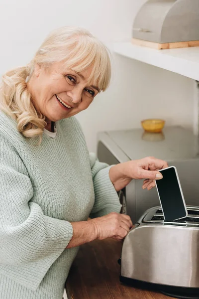 Mulher aposentada alegre com doença de alzheimer colocando smartphone na torradeira — Fotografia de Stock