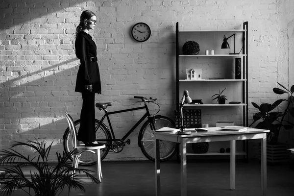 Black and white of attractive woman in black clothes standing on white chair near bike — Stock Photo