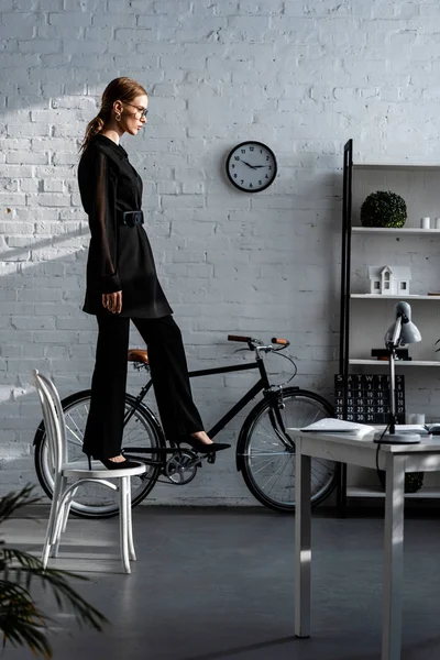 Attraente donna in abiti neri in piedi su sedia bianca vicino al tavolo — Foto stock