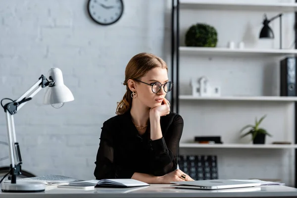 Attractive businesswoman in black clothes and glasses sitting on chair and touching face — Stock Photo