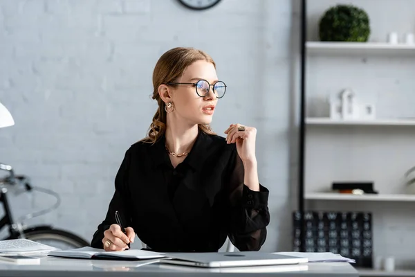 Schöne Geschäftsfrau in schwarzer Kleidung und Brille, die auf einem Stuhl sitzt und Wittling in Notiz nimmt — Stockfoto