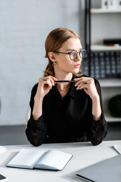 Красива бізнес-леді в чорному одязі і окулярах сидить на стільці і тримає ручку — стокове фото