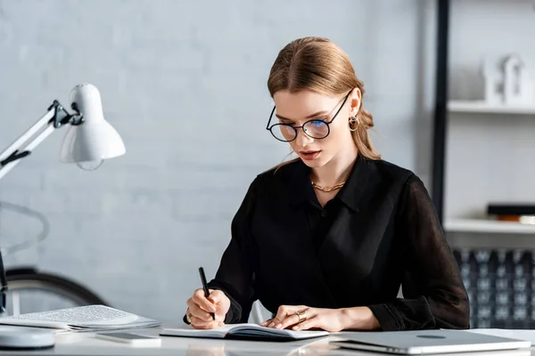 Schöne Geschäftsfrau in schwarzer Kleidung und Brille, die schreibt und auf einem Stuhl sitzt — Stockfoto