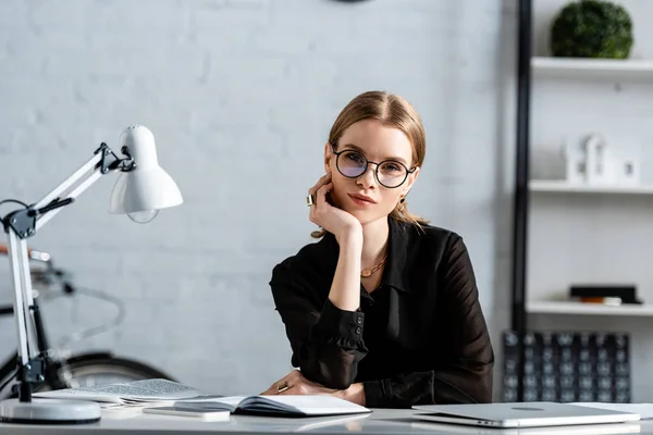 Beautiful businesswoman in black clothes and glasses sitting on chair and holding chin — Stock Photo