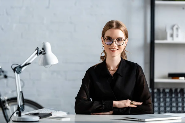 Schöne Geschäftsfrau in schwarzer Kleidung und Brille, die auf einem Stuhl sitzt und in die Kamera lächelt — Stockfoto