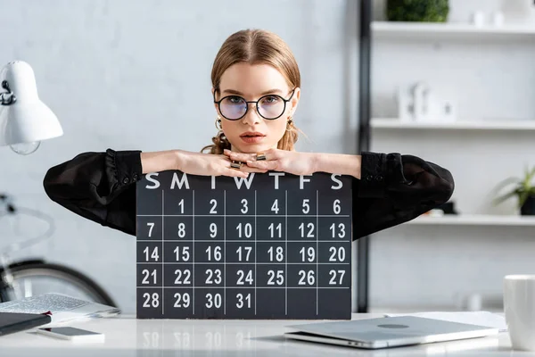 Hermosa mujer de negocios en ropa negra y gafas sentado en la silla y la celebración de un calendario - foto de stock