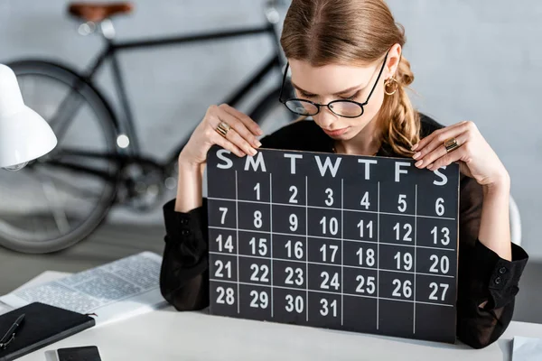 Hermosa mujer de negocios en ropa negra y gafas sentadas en la silla y mirando el calendario - foto de stock