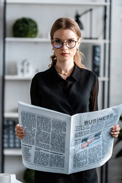 Séduisante femme d'affaires en vêtements noirs et lunettes tenant journal et regardant la caméra — Photo de stock