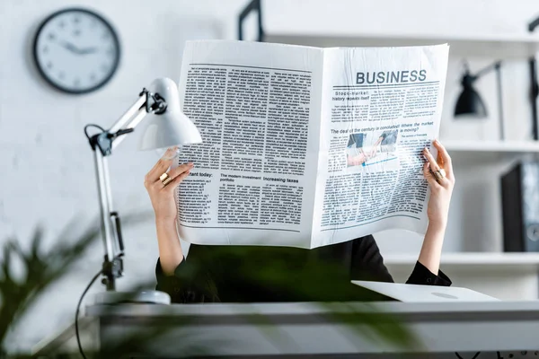 Mujer de negocios en ropa negra sentada en la silla mientras sostiene el periódico de negocios frente a la cara - foto de stock
