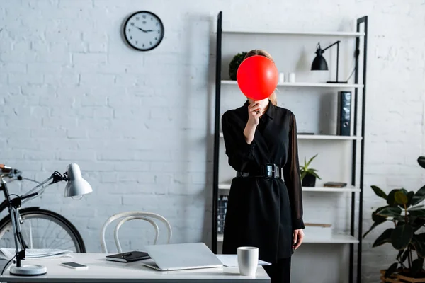 Mujer de negocios en ropa negra de pie cerca de la mesa y escondiendo la cara detrás del globo rojo - foto de stock