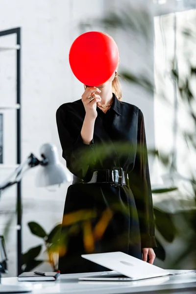 Frau in schwarzer Kleidung steht neben Notizbuch und versteckt Gesicht hinter rotem Luftballon — Stockfoto