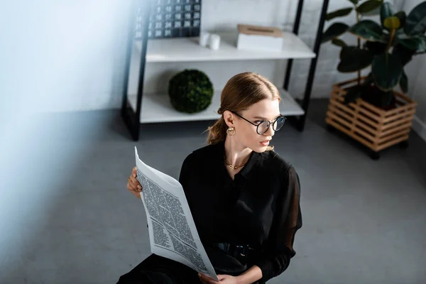 Schöne Geschäftsfrau in schwarzer Kleidung und Brille, die am Tisch sitzt und Zeitung in der Hand hält — Stockfoto