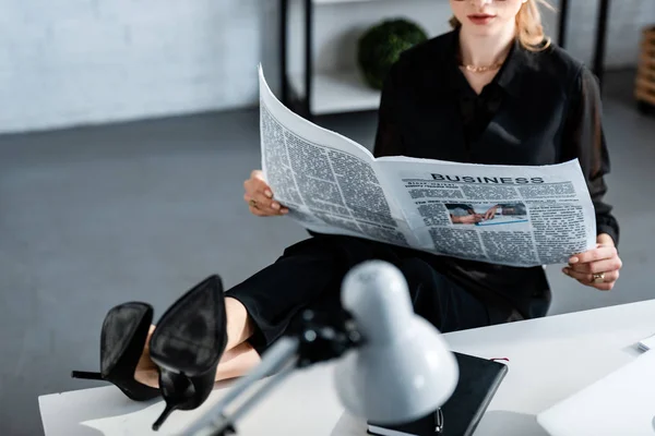 Vista recortada de la mujer de negocios en ropa negra sentada a la mesa y leyendo el periódico - foto de stock