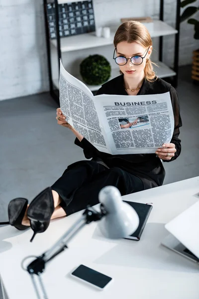 Bella donna d'affari in vestiti neri e scarpe seduti a tavola e tenendo il giornale — Foto stock