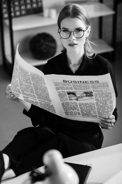 Schwarz-Weiß einer schönen Geschäftsfrau in schwarzen Kleidern und Schuhen, die am Tisch sitzt und die Zeitung in der Hand hält — Stockfoto